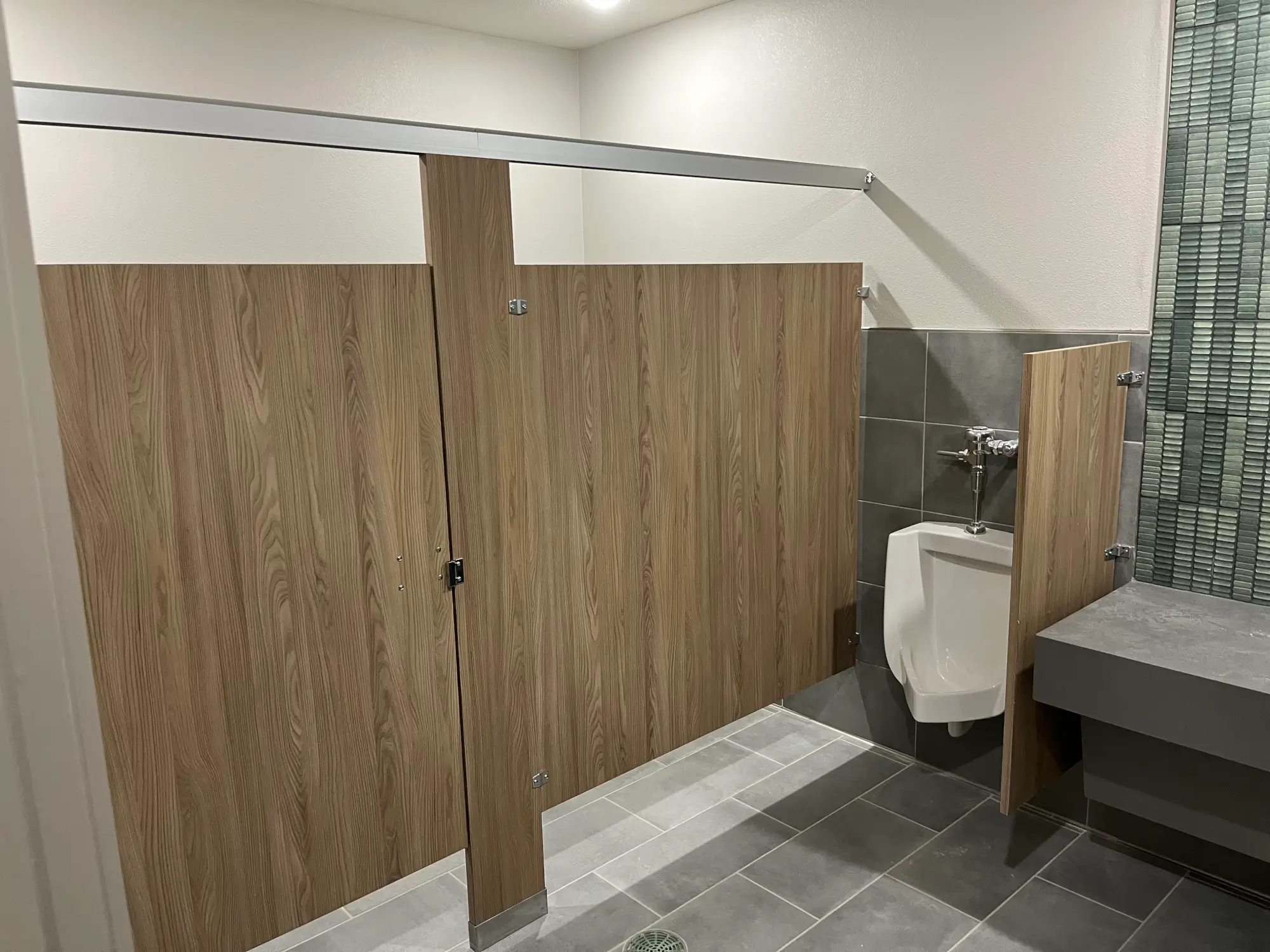 Commercial restroom partitions with wood-look stall doors and urinal privacy screen in a modern bathroom.